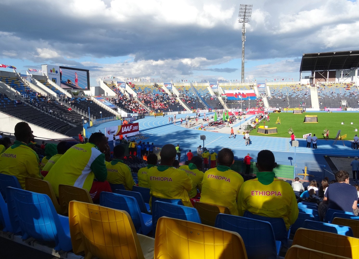 Ein volles Stadion mit Zuschauern bei einem Fußballspiel, mit Bannern, Anzeigetafel, Metalldachträgern und bewölktem Himmel.
