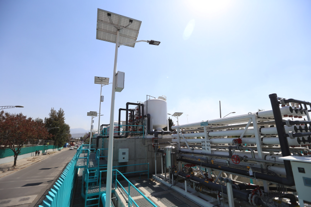 Großes industrielles Gebäude mit ausgedehnten Rohrleitungen und Solarpaneelen auf dem Dach, umgeben von Zäunen, Masten, Schildern, Bäumen und Fahrzeugen, vor Bergen und einem klaren blauen Himmel, Teil einer kalifornischen Wasseraufbereitungsanlage.