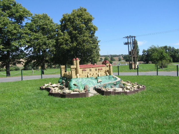 Minigolfplatz mit einem grünen Feld, umgeben von einem Zaun mit Pfählen und Ketten, Bäumen, Strommasten mit Drähten und einem bewölkten Himmel im Hintergrund.