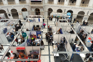 Eine große Gruppe von Menschen auf einer internationalen Fachmesse mit Ständen, Bannern, Tischen, Stühlen und Luftballons in einem Raum mit architektonischen Bögen, Säulen, Geländern und einem Bildschirm im Hintergrund.