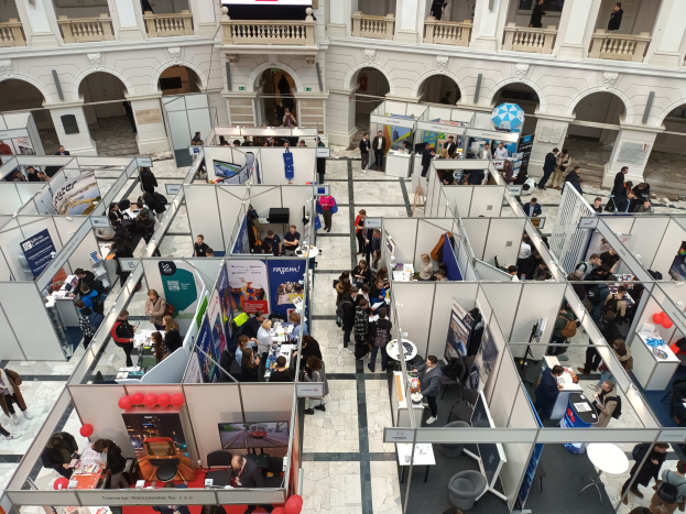 Eine große Gruppe von Menschen auf einer internationalen Fachmesse mit Ständen, Bannern, Tischen, Stühlen und Luftballons in einem Raum mit architektonischen Bögen, Säulen, Geländern und einem Bildschirm im Hintergrund.