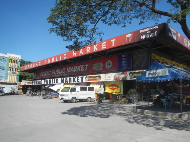 Öffentlicher Markt mit einem vor ihm geparkten Van, umgeben von Gebäuden, Kraftfahrzeugen, Menschen unter einem Zelt, Geländern, Bäumen und einem klaren blauen Himmel.