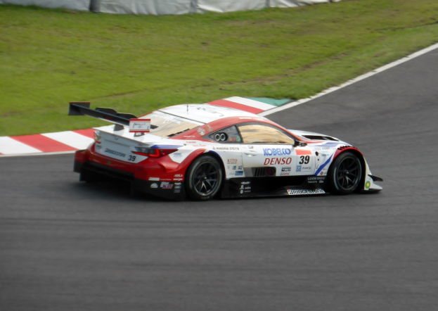 Ein Sportwagen mit Text und Zahlen nimmt an den 24 Stunden von Le Mans teil und fährt auf einer Rennstrecke mit Gras an beiden Seiten und einem Zaun im Hintergrund.