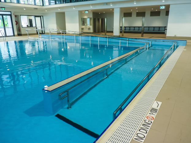 Großes Hallenbad mit Glaswänden, gefliestem Boden und kristallklarem Wasser, umgeben von Säulen, Deckenleuchten und Türen, mit Bäumen im Hintergrund.