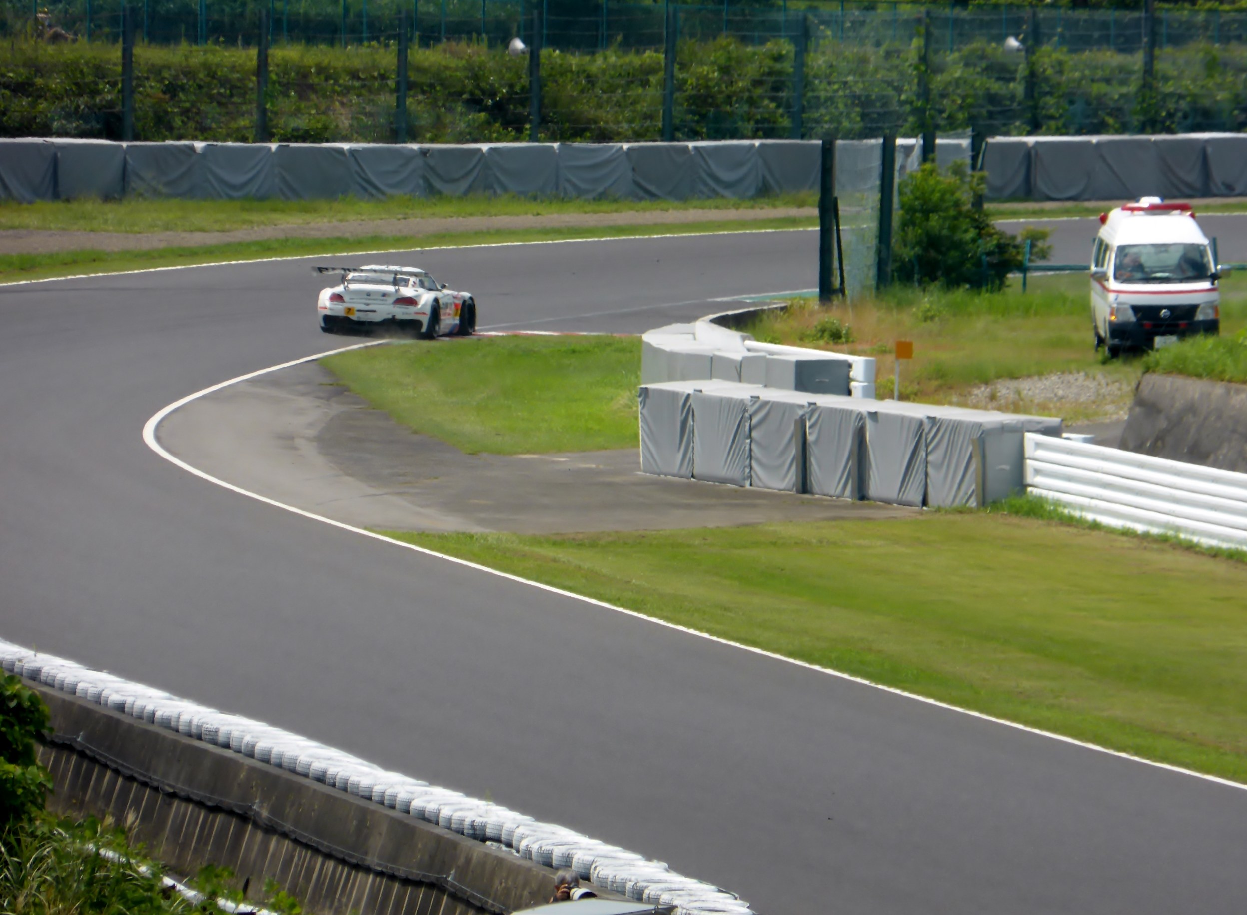 Ein Rennwagen auf einer Rennstrecke mit einem Polizeiwagen im Hintergrund, umgeben von Gras, Pflanzen, Bäumen und einem Zaun, mit einem Fahrzeug auf der rechten Seite und Text unten.