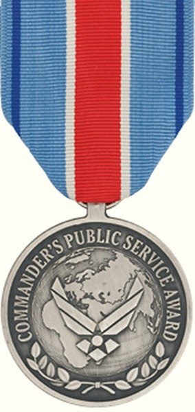 Eine silberne Medaille mit rot-weiß-blauem Band, darauf die Inschrift 'Commander's Public Service Award', vor einem weißen Hintergrund.