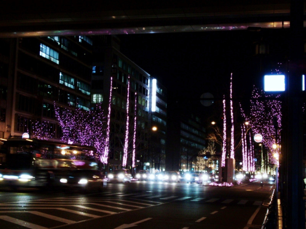 Stadtstraße bei Nacht mit starkem Verkehr, beleuchtet von Straßenlaternen und Weihnachtsbeleuchtung, mit Fahrzeugen, Gebäuden, Laternenmasten und einer Brücke im Hintergrund.
