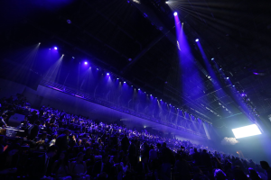 Eine große Menschenmenge sitzt einer Bühne mit Deckenbeleuchtung gegenüber, mit einem Bildschirm auf der rechten Seite.