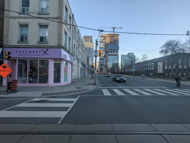 Stadtstraße mit fahrenden Autos, ein Fahrradfahrer auf dem Gehweg, Verkehrsampeln, Schilder, Gebäude, Bäume und Himmel.