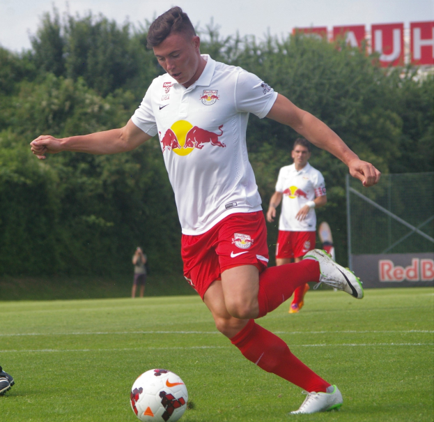 Ein Fussballer in Uniform kickt einen Ball auf einem grünen Feld mit Bäumen und einem klaren blauen Himmel im Hintergrund, wobei ein "RB Leipzig"-Schild sichtbar ist.