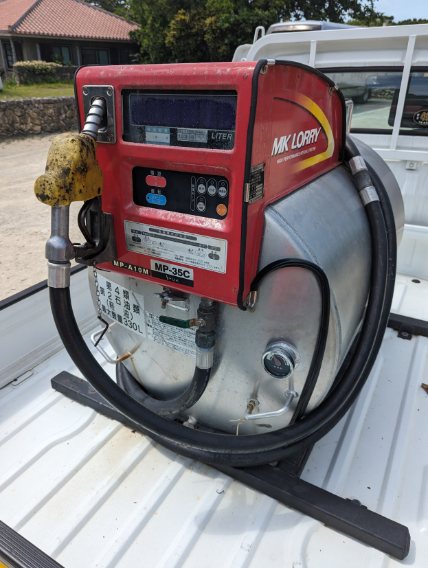 Ein Lastwagen mit einer Gaspumpe am Heck, umgeben von Bäumen, einem Haus und einem klaren blauen Himmel.