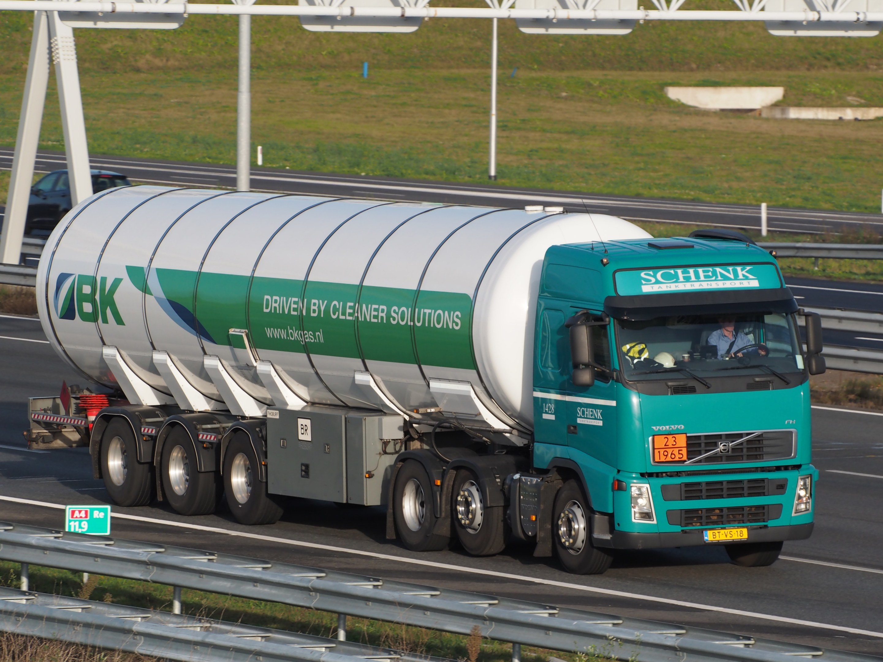Ein großer Tanklastzug fährt auf einer Autobahn mit einer Person am Steuer, umgeben von Leitplanken und Gras, mit Schildern im Hintergrund.