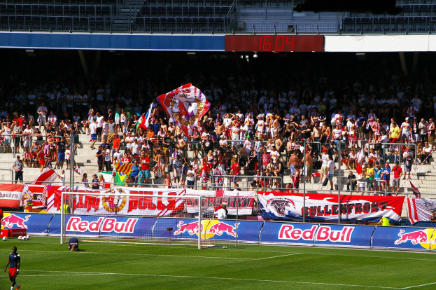 Ein Fußballspiel in einem Stadion mit Spielern auf dem Feld, einem Torpfosten mit Netz, Bannern, einem Metallzaun, einer Anzeigetafel, einem Bildschirm und einem Dach mit Deckenbeleuchtung.