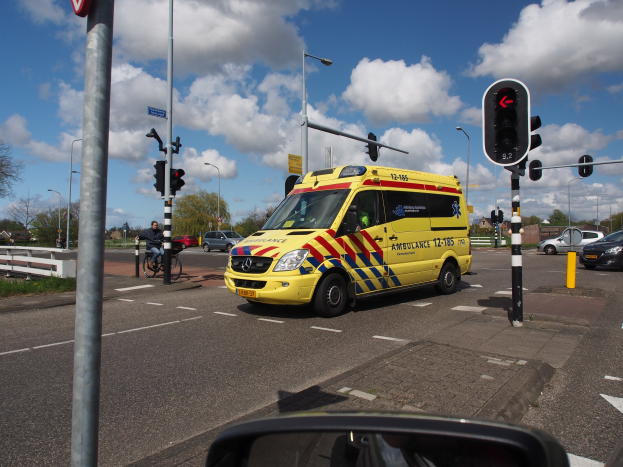 Ein Rettungswagen fährt auf einer Straße neben einer Ampel, mit einem Radfahrer im Vordergrund, umgeben von Laternenmasten, Bäumen, Gebäuden und einem bewölkten Himmel, mit einem Spiegel unten.