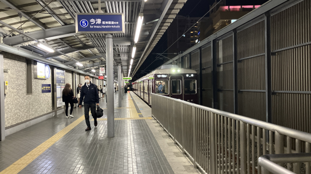 Zug einfährt auf einem Bahnsteig mit Menschen in der Nähe, einem Geländer auf der rechten Seite, Gebäuden im Hintergrund, Bahnsteiglampen und einer Anzeigetafel mit Text.