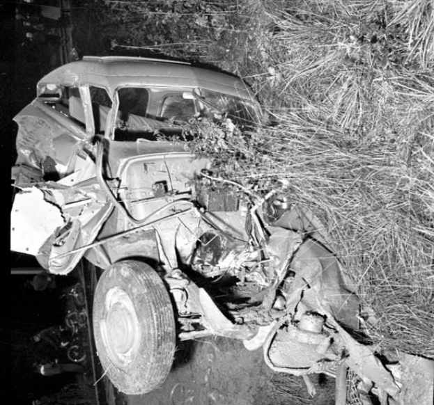 Ein Schwarz-Weiß-Foto eines schwerbeschädigten Autos am Straßenrand, mit einer eingedrückten Front, zersplitterten Fenstern, einem Baum im Hintergrund und verstreuten Gegenständen an der Unfallstelle.