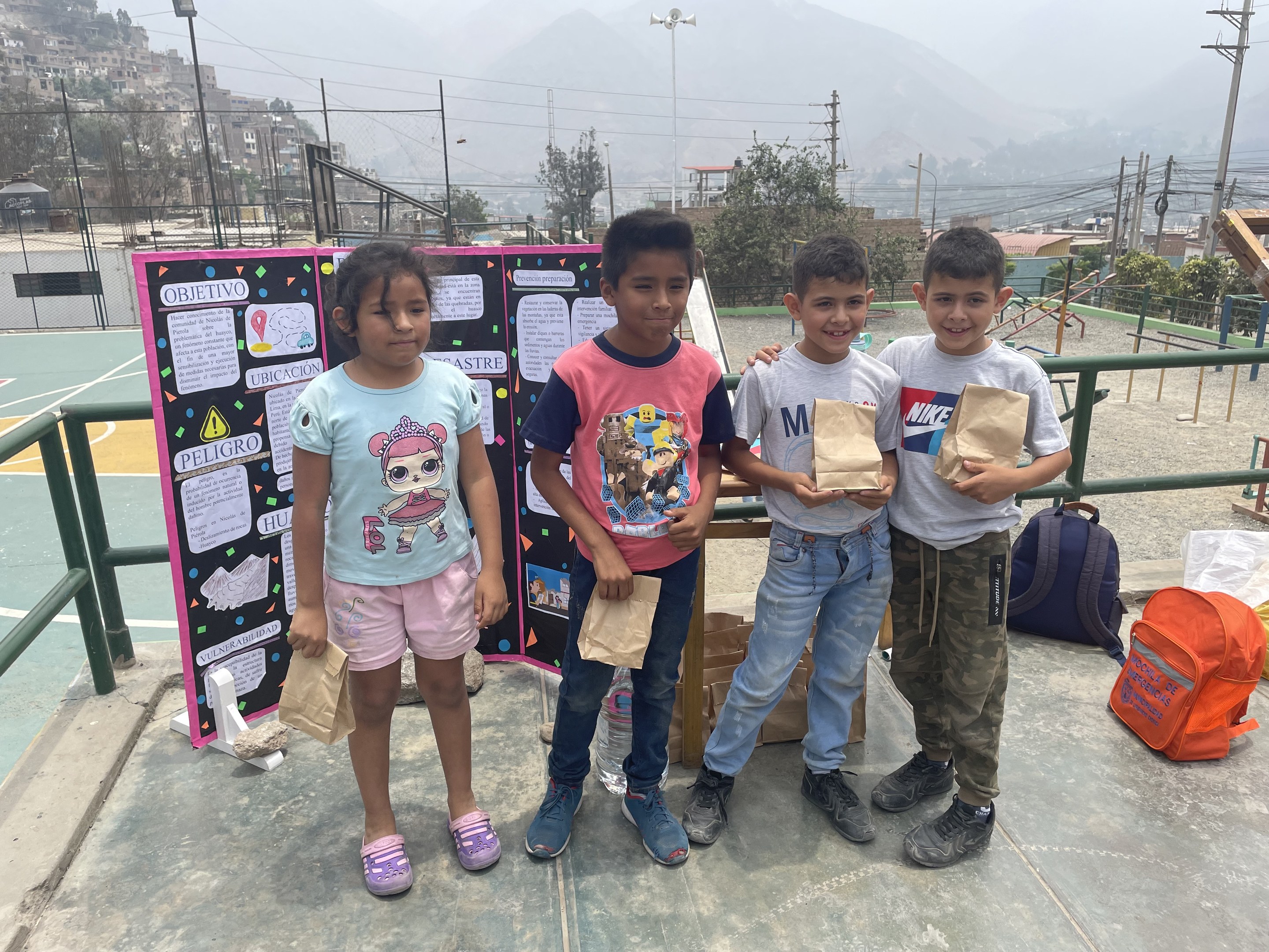 Gruppe von Kindern auf einem Basketballfeld mit Papiertüten in den Händen, mit Tüten und Gegenständen rechts und einer Tafel mit Text links, vor einem Hintergrund aus Zäunen, Masten, Straßenlaternen, Bäumen, Gebäuden, Höhen und Himmel.