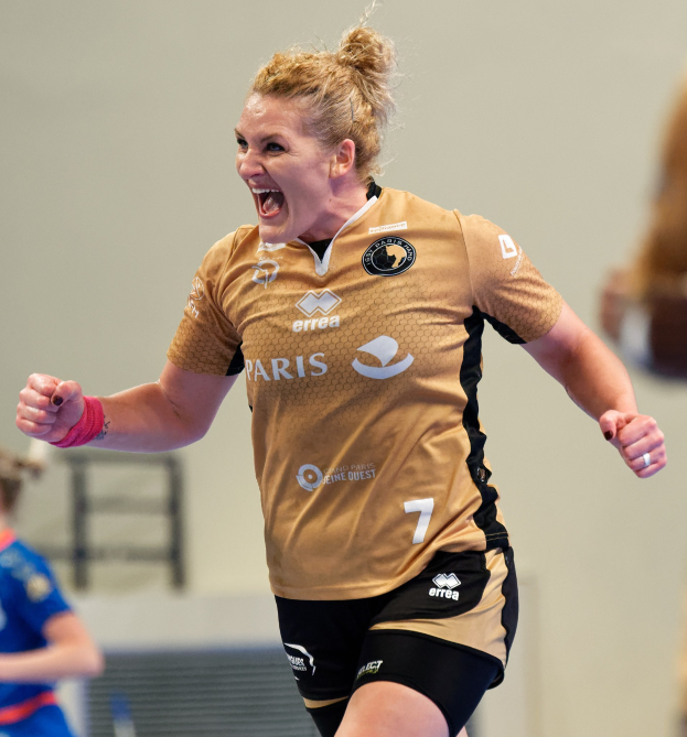 Eine Frau in einem braunen Hemd und schwarzen Shorts, die auf einem Futsalplatz läuft und feiert, mit zwei anderen Frauen im Hintergrund.