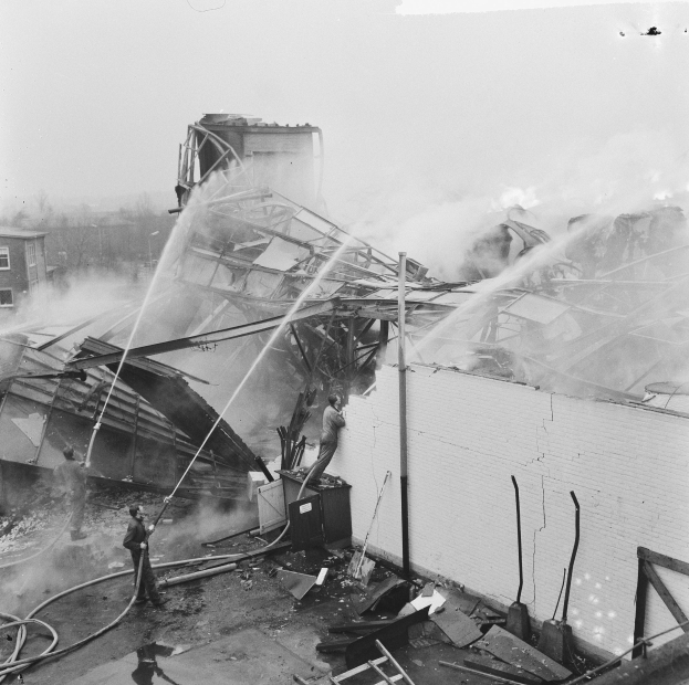 Schwarzes und weißes Foto von Feuerwehrleuten auf einem Dach, die ein Feuer löschen, während Menschen unten mit Leitern und Rohren helfen.