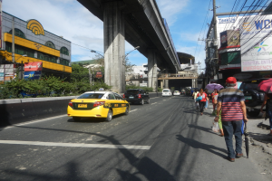 Gelbes Taxi fährt eine Stadtstraße mit hohen Gebäuden, Fußgängern auf dem Gehweg mit Regenschirmen, einer Brücke im Hintergrund, bewölktem Himmel und Oberleitungen.