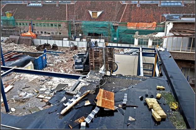 Baustelle mit einem Kran, umgeben von Gebäuden, Bäumen, Absperrungen und anderen Gegenständen, mit Text am unteren Rand.