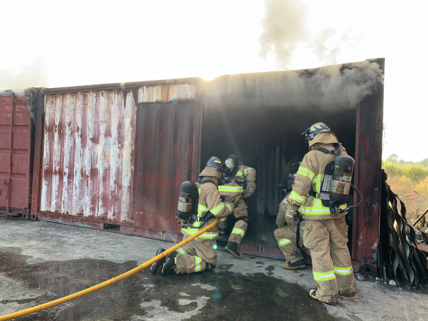 Feuerwehrleute in Schutzausrüstung löschen ein Feuer in einem Seecontainer, mit Rauchentwicklung und Grünzeug im Hintergrund.