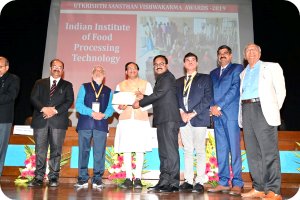 Eine Gruppe von Männern auf einer Bühne mit einem Zertifikat und einem Bildschirm im Hintergrund mit der Aufschrift "Indian Institute of Food Processing Technology Awards 2019" und Blumenschmuck.