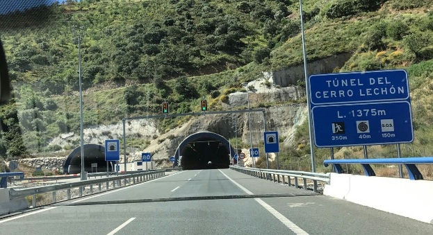 Auto fährt durch einen Tunnel neben einer Straße mit Geländern, Schildern, Pfosten, Verkehrszeichen, Bäumen, Felsen und einem klaren blauen Himmel.