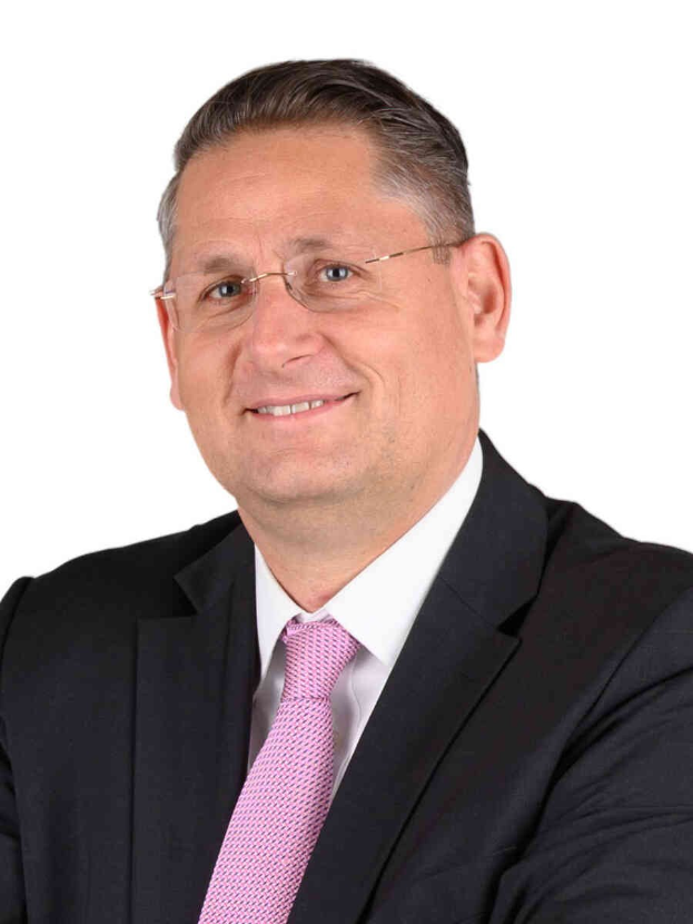 Ein lächelnder Mann im Anzug, mit Krawatte und Brille vor einem weißen Hintergrund.