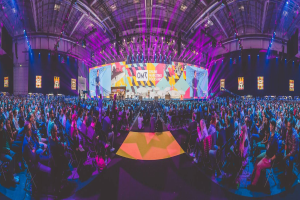 Eine große Menschenmenge, die auf Stühlen sitzt und einer hell erleuchteten Bühne mit mehreren Personen darauf gegenübersitzt, umgeben von Bildschirmen, wahrscheinlich bei einem Online-Marketing-Event.