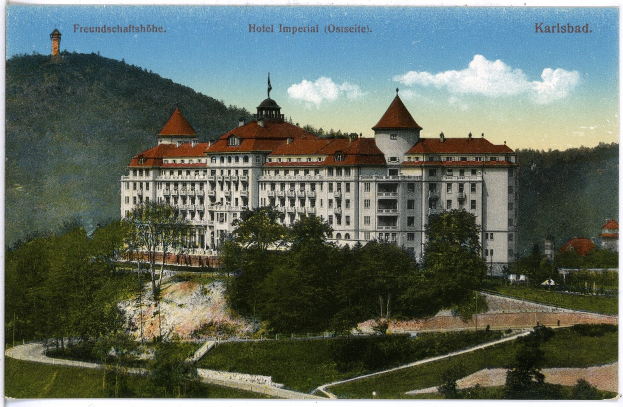 Eine alte Postkarte zeigt das Hotel Imperial in Karlsbad, Deutschland, einen großen Bau mit zahlreichen Fenstern, umgeben von Bäumen, Pflanzen und Gras, mit einem Weg im Vordergrund und Hügeln und einem bewölkten Himmel im Hintergrund sowie Text oben auf dem Bild.