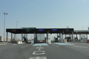 Eine Mautstelle am Straßenrand mit einem vor ihr geparkten Lkw, weiße und blaue Fahrbahnmarkierungen, Laternenmäste, Schilder und andere Gegenstände auf beiden Seiten, mit Gebäuden, Bäumen und einem klaren blauen Himmel im Hintergrund.