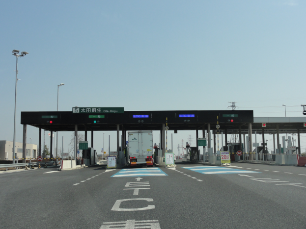 Eine Mautstelle am Straßenrand mit einem vor ihr geparkten Lkw, weiße und blaue Fahrbahnmarkierungen, Laternenmäste, Schilder und andere Gegenstände auf beiden Seiten, mit Gebäuden, Bäumen und einem klaren blauen Himmel im Hintergrund.