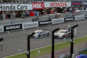 Eine Gruppe von Autos, die auf einer Rennstrecke fahren, umgeben von einem Metallzaun, Gras, Schildern, Metallpfosten, einer Barriere, einem weiteren Zaun, einem Gebäude mit Fenstern und einem Dach mit Deckenbeleuchtung.