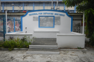 Ein Denkmal für verstorbene Brüder steht vor einem Gebäude mit einem blauen und weißen Schild, Fenstern, einem Metallzaun, Pflanzen, einer Treppe, Pfählen, Drähten und einem Baum unter einem bewölkten Himmel.