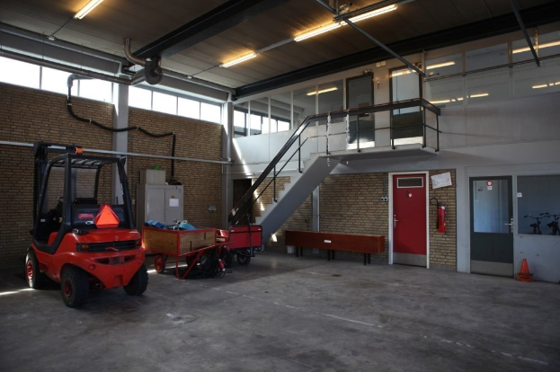 Ein großes Lagerinneres mit einem roten Gabelstapler, einem Wagen mit Gegenständen, einer Bank, einem Feuerlöscher, einer Treppe mit Geländern, Türen, Fenstern, Deckenleuchten, Rohren und einem sichtbaren Rad.
