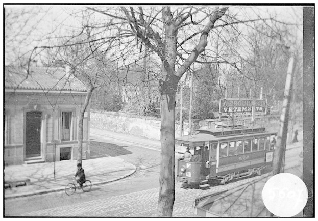 Ein Schwarz-Weiß-Foto einer Straßenbahn auf einer Stadtstraße, mit einer Person, die ein Fahrrad vor ihr fährt, einigen Menschen in der Straßenbahn und Bäumen und einem Gebäude im Hintergrund.