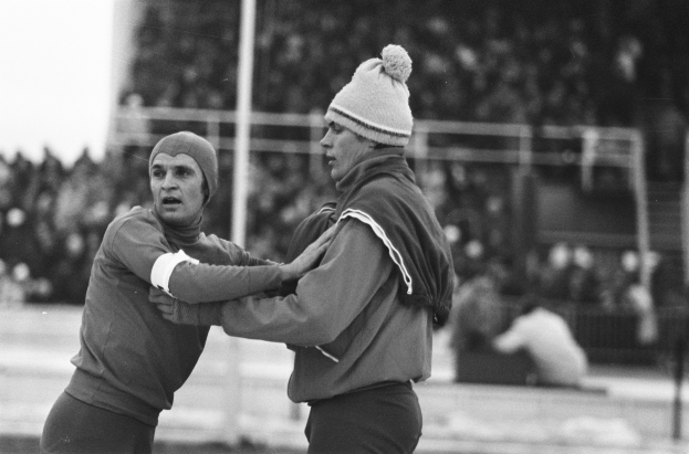 Zwei Männer in Jacken und Mützen kämpfen auf einem Feld mit unscharfen Zuschauern, Pfählen und einem Zaun im Hintergrund.