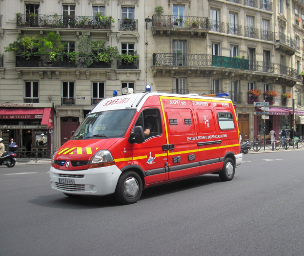 Rettungswagen auf der Straße neben einem Gebäude mit Fenstern, Geländern, Pflanzen und anderen Gegenständen geparkt, mit ein paar Menschen und Fahrrädern im Hintergrund.