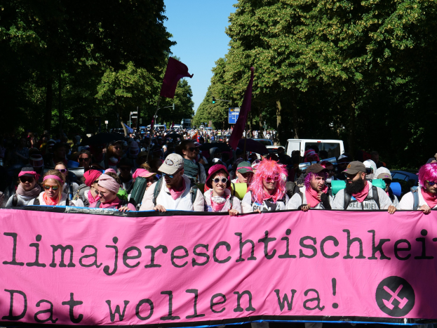 Gruppe von Menschen marschiert die Straße entlang und hält eine pinkfarbene "Women's March in Berlin"-Fahne, einige tragen Mützen und Schutzbrillen, andere halten Fahnen, mit Bäumen, Fahrzeugen und Himmel im Hintergrund.