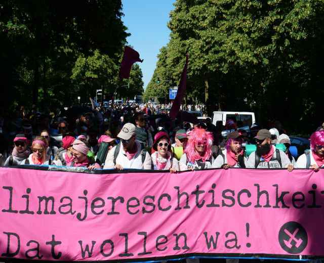 Gruppe von Menschen marschiert die Straße entlang und hält eine pinkfarbene "Women's March in Berlin"-Fahne, einige tragen Mützen und Schutzbrillen, andere halten Fahnen, mit Bäumen, Fahrzeugen und Himmel im Hintergrund.