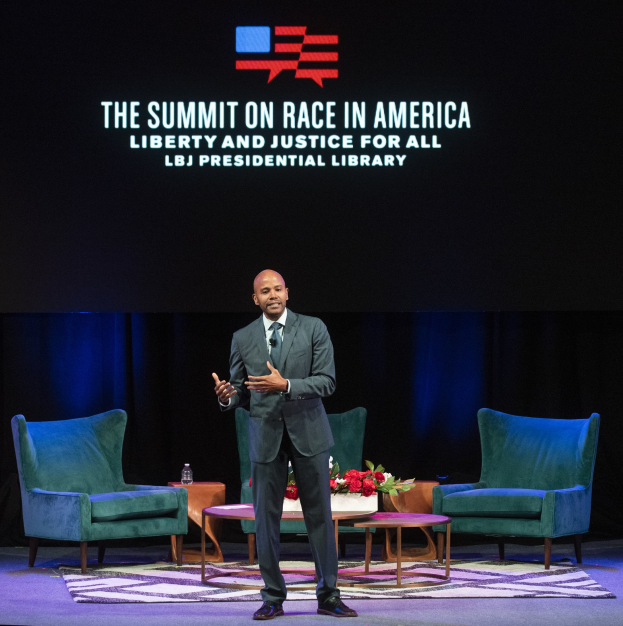 Ein Mann in einem Blazer, Krawatte und Hemd steht auf einer Bühne und spricht bei der Veranstaltung "The Summit on Race in America: Liberty and Justice for All" in der LBJ Presidential Library. Hinter ihm befinden sich zwei Stühle und ein Tisch mit einem Blumenstrauß und einer Flasche darauf. Im Hintergrund ist ein Vorhang und ein Bildschirm mit der Aufschrift "The Summit on Race in America: Liberty and Justice for All" zu sehen.