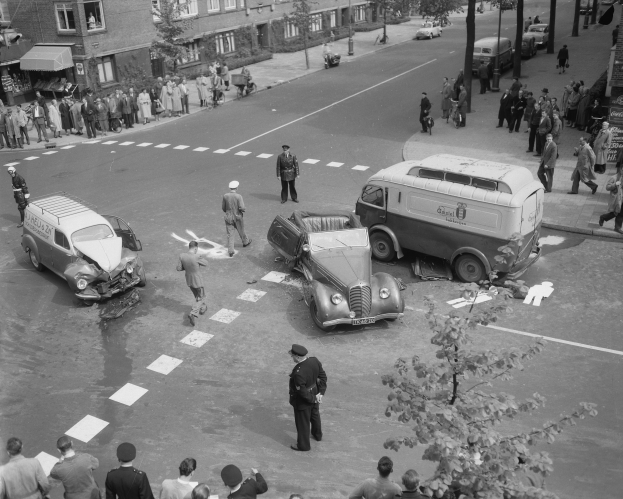 Schwarz-weiß-Szene eines Autounfalls an einer Straßenkreuzung mit mehreren Fahrzeugen, darunter ein Lieferwagen, und einer Gruppe von Menschen, die danebenstehen, mit Gebäuden, Bäumen, Pfählen und Texttafeln im Hintergrund.