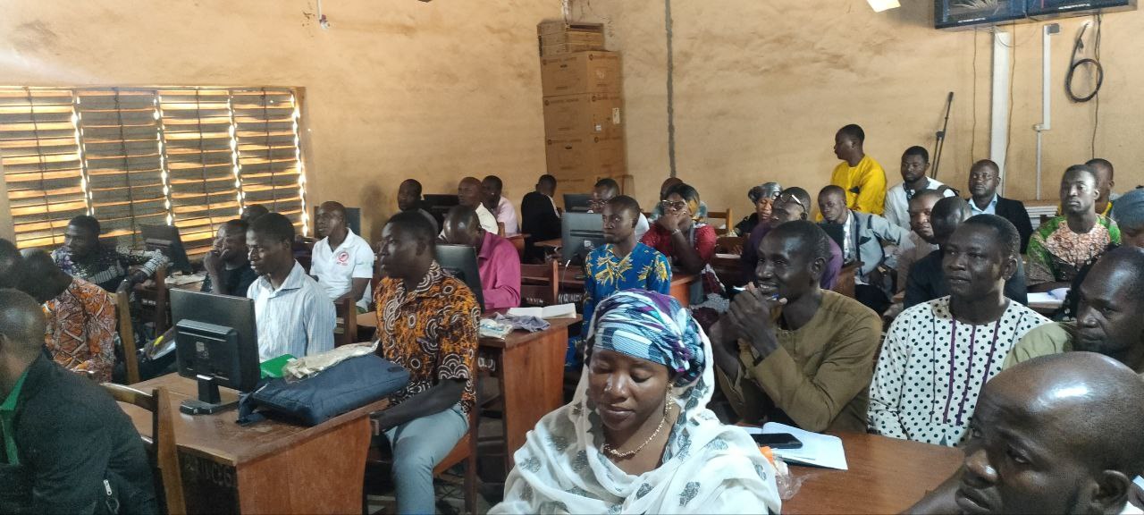 Eine Gruppe von Menschen, die an Tischen in einem Klassenzimmer mit Monitoren, Taschen, Büchern und Papieren sitzen und an einer Techniktraining teilnehmen; ein Fenster auf der linken Seite, Kartons und ein Fernseher an der Wand im Hintergrund.