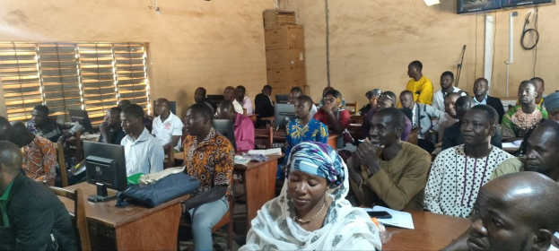 Eine Gruppe von Menschen, die an Tischen in einem Klassenzimmer mit Monitoren, Taschen, Büchern und Papieren sitzen und an einer Techniktraining teilnehmen; ein Fenster auf der linken Seite, Kartons und ein Fernseher an der Wand im Hintergrund.