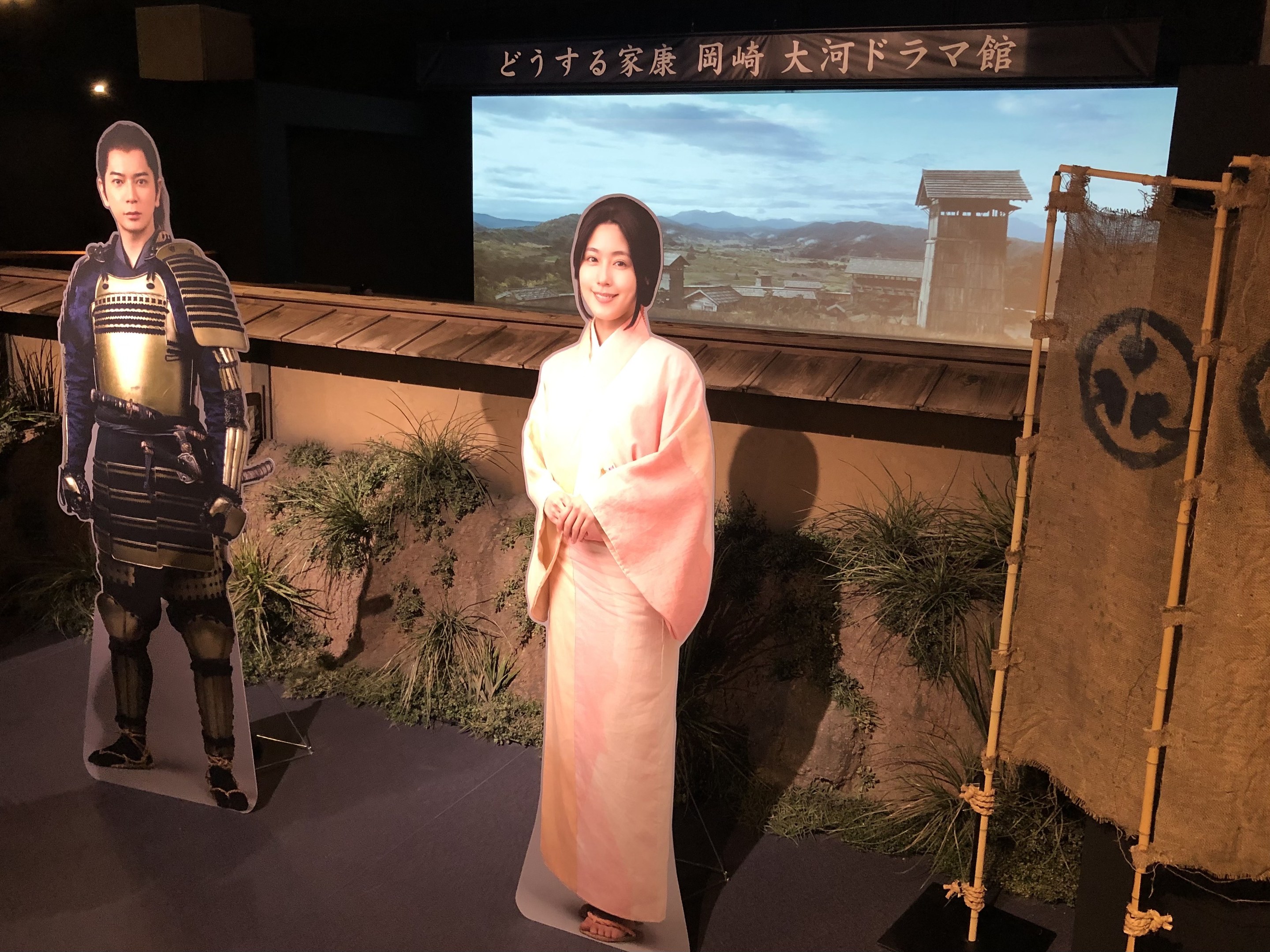 Eine Frau in einem rosa Kimono und ein Mann in einem Samurai-Kostüm, die gemeinsam in einer Szene aus einem japanischen Film lächeln, mit Pflanzen, einem Bildschirm und Lichtern im Hintergrund.