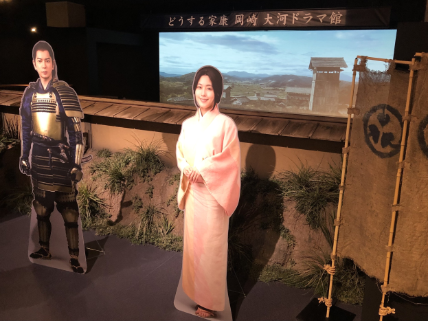 Eine Frau in einem rosa Kimono und ein Mann in einem Samurai-Kostüm, die gemeinsam in einer Szene aus einem japanischen Film lächeln, mit Pflanzen, einem Bildschirm und Lichtern im Hintergrund.