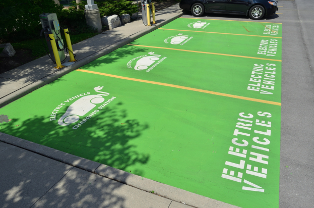 Elektroauto an einer Ladesäule geparkt, markiert durch eine grüne Bodenmatte mit Text und Bildern in einer von Bäumen und einem Gebäude umgebenen Parkfläche.