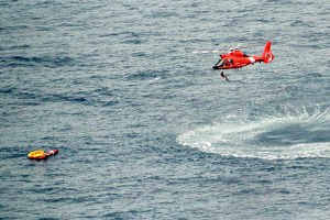 Roter Hubschrauber über tiefblauem Meer mit einem Rettungsboot darunter, Schwimmwesten an der Seite des Bootes, strahlender Sonnenschein auf dem Wasser.