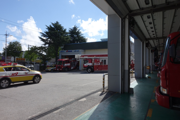 Feuerwache mit mehreren Feuerwehrautos vorne geparkt, ein Schuppen rechts und im Hintergrund Bäume, Gebäude, Strommäste mit Drähten und ein bewölkter Himmel.